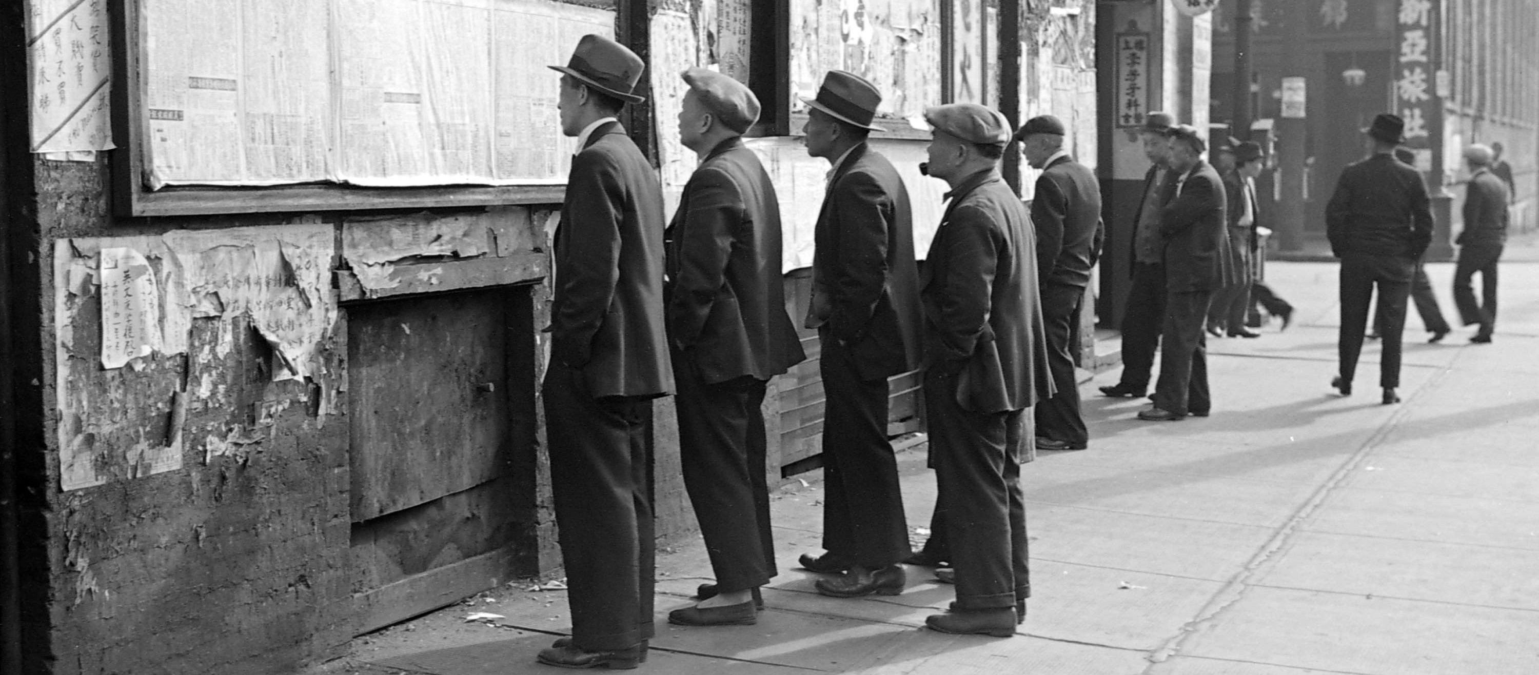 A group of men stand on the sidewalk in Vancouver's Chinatown, reading newspapers posted to a board on the side of a building.