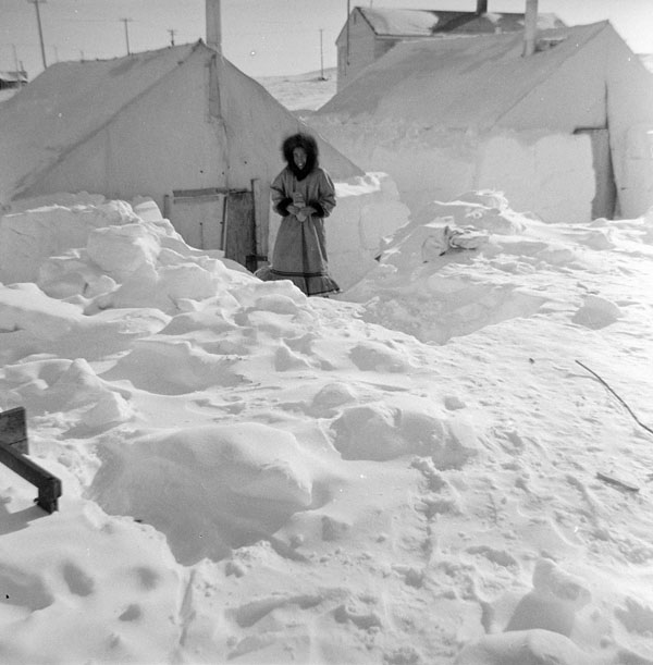 Une femme inuite se tient devant deux tentes entourées de bancs de neige.