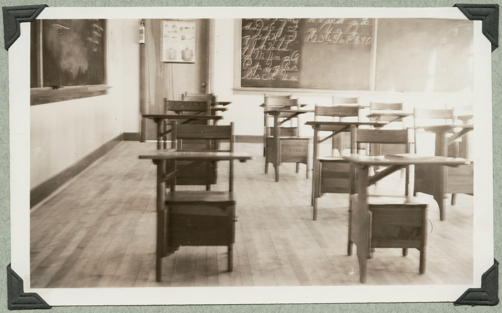 An empty classroom with chairs attached to desktops and blackboards on the walls. One of the blackboards has the alphabet and the numbers 0-9 written on it.